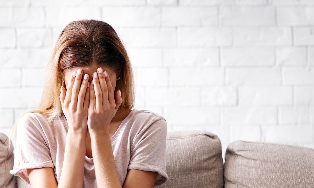A woman sits on a couch, covering her face with her hands, conveying a sense of distress or contemplation.
